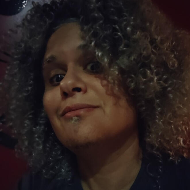 Facsimile Hair Mixed Boricua person with gray curly hair and glasses, black shirt and a yellow background.