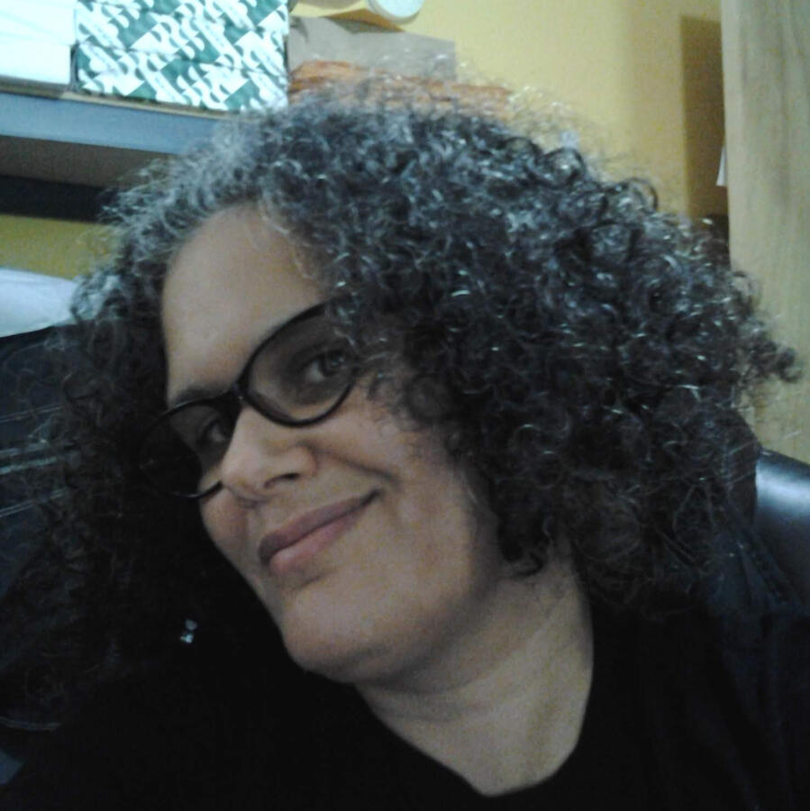 Mixed Boricua person with black curly hair and glasses, black shirt and a yellow background.