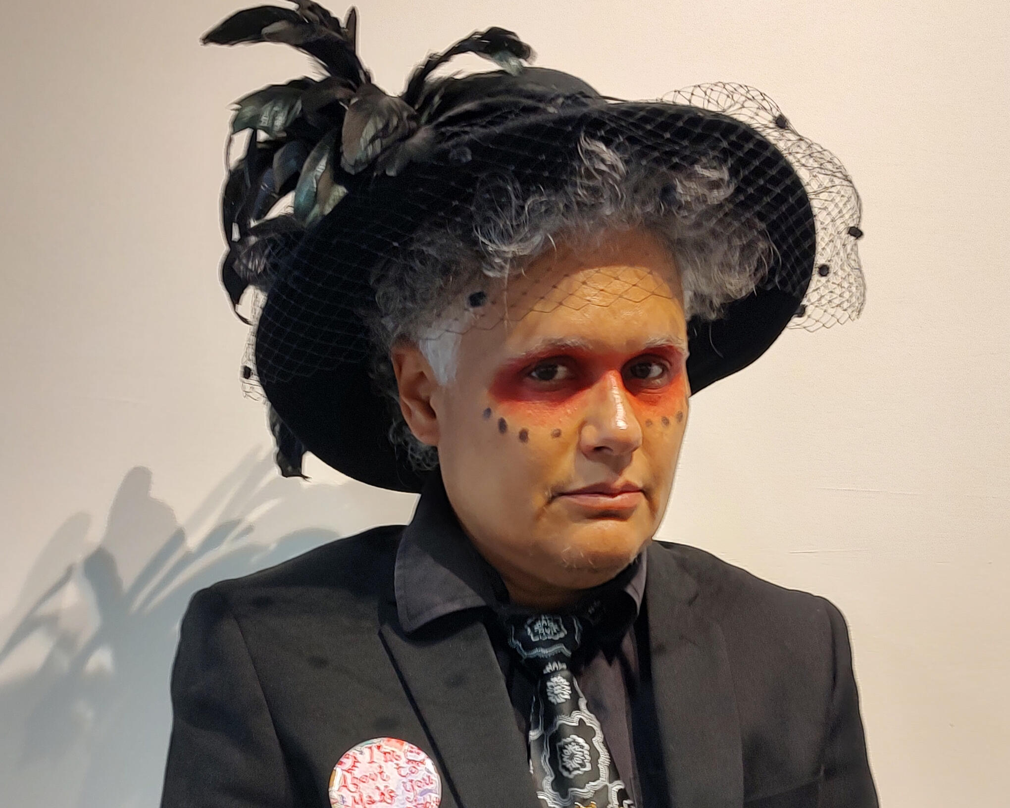 Tanamá Kao Tanamá Kao wearing a black suit and a fanciful black hat with feathers against a white background.