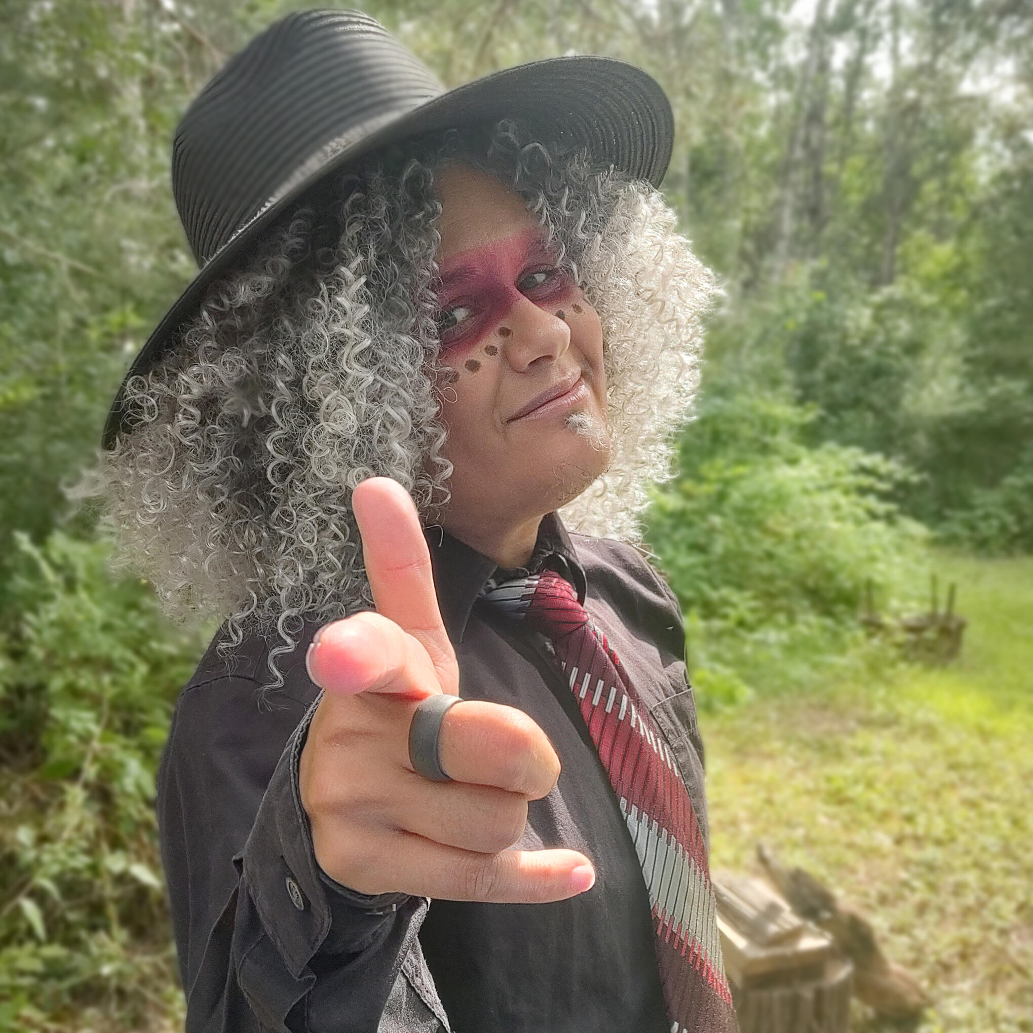 Tanamá Kao, a gray-haired person with coily curls is standing in front of a forest background.