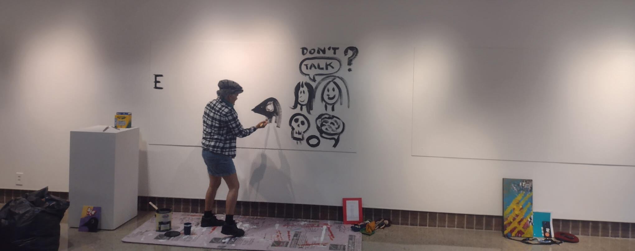 Tanamá dressed in flannel, shorts, a news boy hat, and shoes, painting the Color Me Safe wall at Tanamá Kao's Battle For Safety